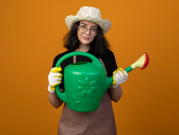 Erfreute junge brünette Gärtnerin in optischen Gläsern und in Uniform mit Gartenhut und Handschuhen hält Gießkanne isoliert auf orange Wand