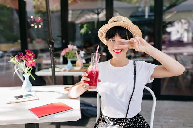 Erfreute junge Frau im Strohhut, die mit Friedenszeichen aufwirft und Glas des kalten Getränks im Sommermorgen hält