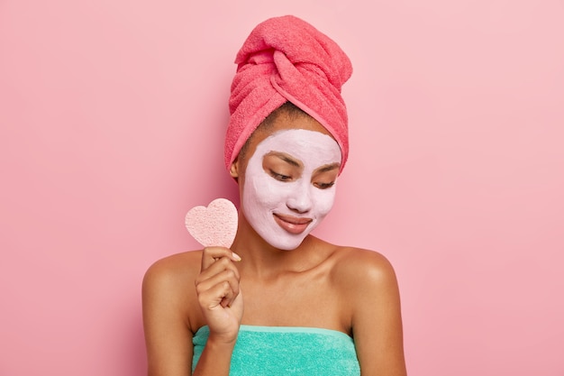 Erfreute junge Frau konzentriert sich nach unten, trägt Gesichtsmaske aus Ton, hält kosmetischen Schwamm zum Entfernen von Make-up, zeigt nackte Schultern, eingewickelt in Badetuch, isoliert auf rosa Studiowand