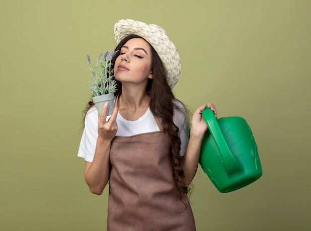 Erfreute junge Gärtnerin in Uniform mit Gartenhut hält Gießkanne und schnüffelt Blumen im Blumentopf isoliert auf olivgrüner Wand