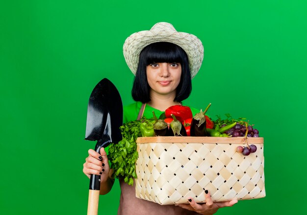 Erfreute junge gärtnerin in uniform mit gartenhut hält spaten und gemüsekorb