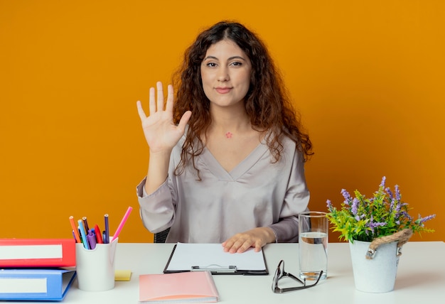 Erfreute junge hübsche Büroangestellte, die am Schreibtisch mit Bürowerkzeugen sitzt, die Hallo-Geste lokalisiert auf Orange zeigen