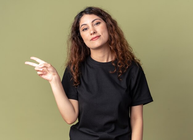Erfreute junge hübsche Frau, die nach vorne schaut und das Friedenszeichen isoliert auf olivgrüner Wand tut?