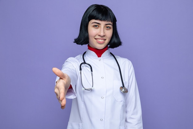 Erfreute junge hübsche kaukasische Frau in Arztuniform mit Stethoskop, die die Hand aushält