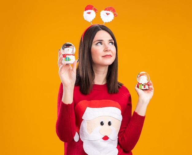 Erfreute junge hübsche kaukasische mädchen mit weihnachtsmann-stirnband und pullover mit schneemann und weihnachtsmann-figuren, die einzeln auf orangefarbenem hintergrund mit kopierraum nachschlagen