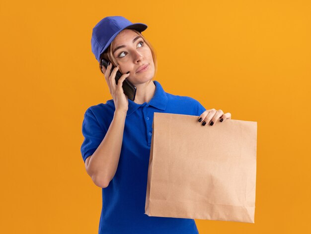 Erfreute junge hübsche Lieferfrau in Uniform hält Papierpaket und spricht am Telefon, das Seite betrachtet, die auf orange Wand lokalisiert ist