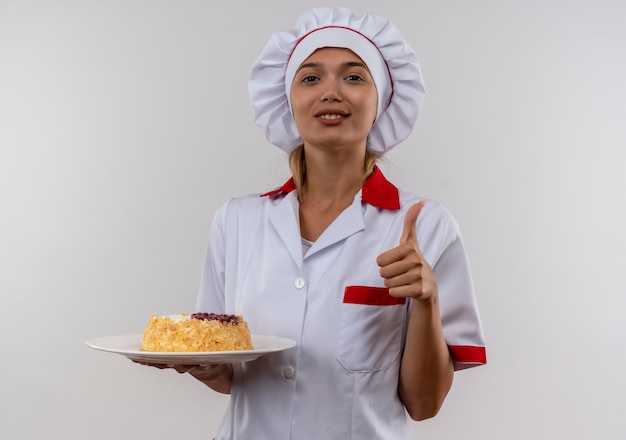 Erfreute junge köchin, die kochuniform trägt, die kuchen auf platte ihren daumen oben auf isolierter weißer wand mit kopienraum hält