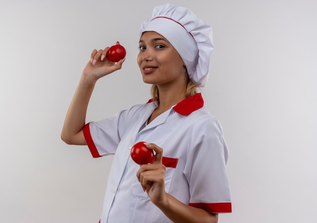 Erfreute junge Köchin, die Kochuniform trägt, die Tomates auf isolierter weißer Wand hält