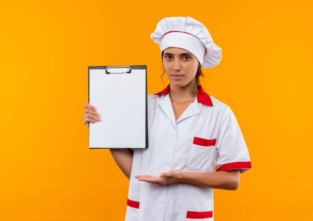 Erfreute junge Köchin, die Kochuniform trägt und mit Hand Zwischenablage auf isolierter gelber Wand mit Kopienraum zeigt