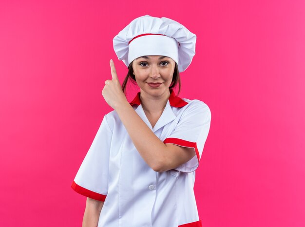 Erfreute junge Köchin, die Kochuniform trägt, zeigt nach oben isoliert auf rosa Hintergrund