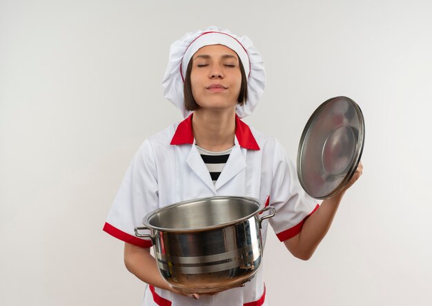 Erfreute junge Köchin im einheitlichen Öffnungsdeckel des Topfes des Kochs mit geschlossenen Augen lokalisiert auf weißer Wand