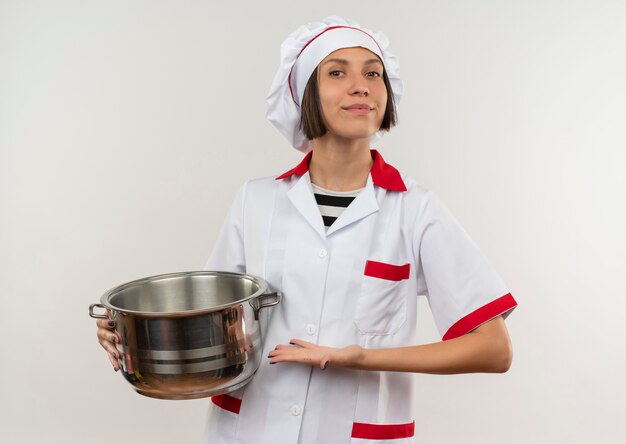 Erfreute junge Köchin in der Kochuniform, die auf weißem Hintergrund lokalisiert und auf Topf zeigt