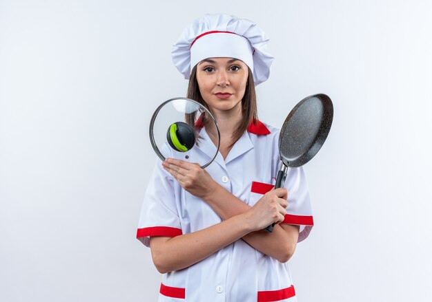 Erfreute junge Köchin in Kochuniform, die Bratpfanne mit Deckel auf weißem Hintergrund hält und überquert