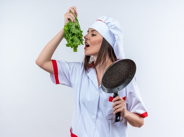 erfreute junge Köchin in Kochuniform, die Salat mit Bratpfanne isoliert auf weißer Wand hält