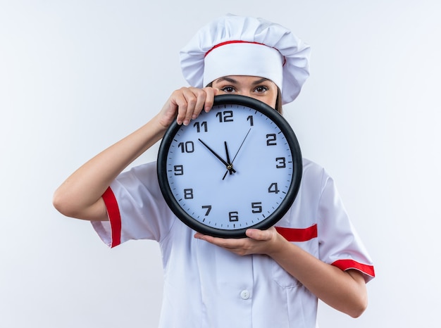 erfreute junge Köchin mit Kochuniform bedecktes Gesicht mit Wanduhr isoliert auf weißer Wand