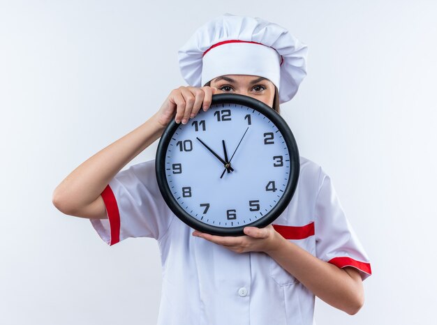 erfreute junge Köchin mit Kochuniform bedecktes Gesicht mit Wanduhr isoliert auf weißer Wand