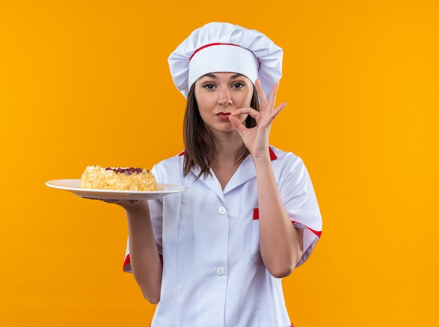 erfreute junge Köchin mit Kochuniform, die Kuchen auf dem Teller hält und eine köstliche Geste zeigt, die auf orangefarbener Wand isoliert ist?