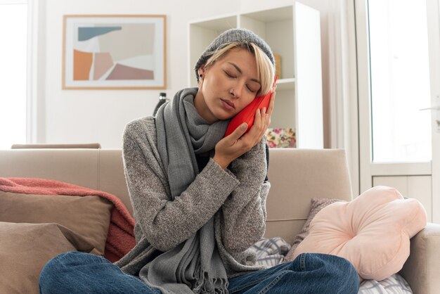 erfreute junge kranke slawische Frau mit Schal um den Hals, die eine Wintermütze trägt und den Kopf auf eine Wärmflasche setzt, die auf der Couch im Wohnzimmer sitzt