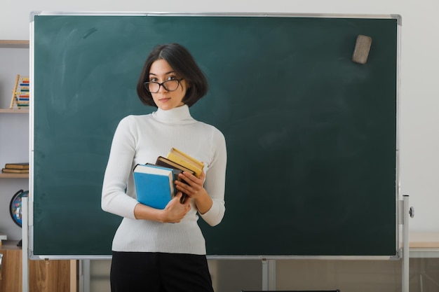 erfreute junge Lehrerin, die Bücher hält, die vor der Tafel im Klassenzimmer stehen