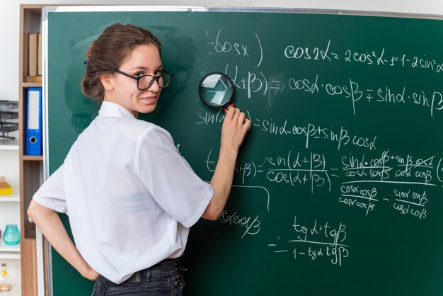 Erfreute junge Mathematiklehrerin mit Brille, die hinter der Tafel vor der Tafel steht standing