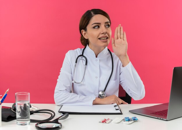 Erfreute junge Ärztin, die medizinische Robe und Stethoskop trägt, die am Schreibtisch mit medizinischen Werkzeugen und Laptop sitzt, die Seite betrachten, die Hand in Luft hält