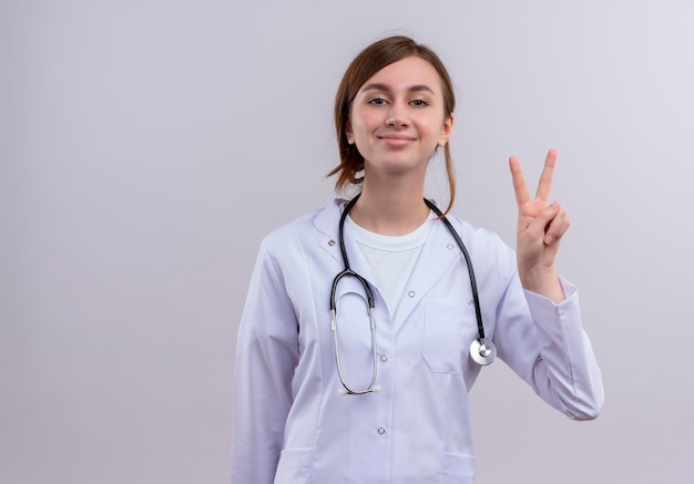 Erfreute junge Ärztin, die medizinische Robe und Stethoskop trägt, die Friedenszeichen mit Kopienraum tun
