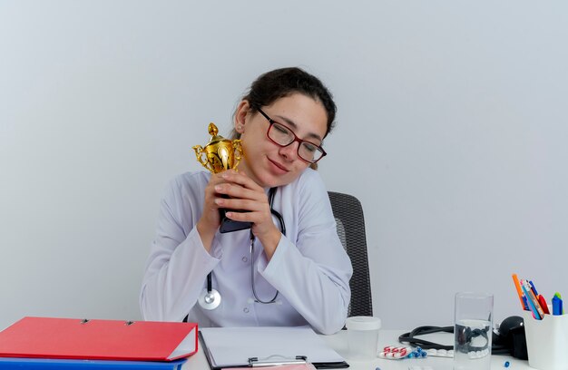 Erfreute junge Ärztin, die medizinische Robe und Stethoskop und Brille trägt, die am Schreibtisch mit medizinischen Werkzeugen sitzen, die Gewinnerbecher mit geschlossenen Augen lokalisiert halten