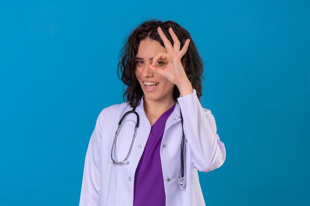 Erfreute junge Ärztin, die weißen Kittel mit Stethoskop trägt, das fröhlich lächelt und ok Zeichen macht, das durch dieses Zeichen schaut, das auf lokalisiertem Blau steht