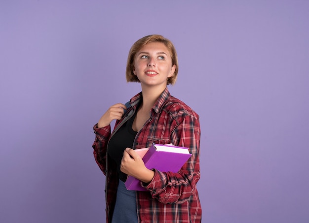 Erfreute junge slawische Studentin mit Rucksack steht seitlich und hält Buch und Notizbuch