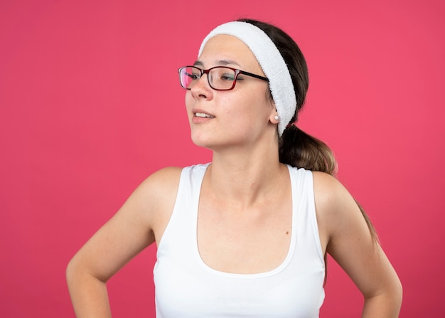 Erfreute junge sportliche Frau in der optischen Brille, die Stirnband und Armbänder trägt, schaut auf Seite, die auf rosa Wand lokalisiert wird