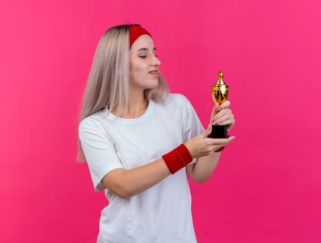 Erfreute junge sportliche Frau mit Hosenträgern, die Stirnband und Armbänder tragen, hält und betrachtet Gewinnerpokal lokalisiert auf rosa Wand