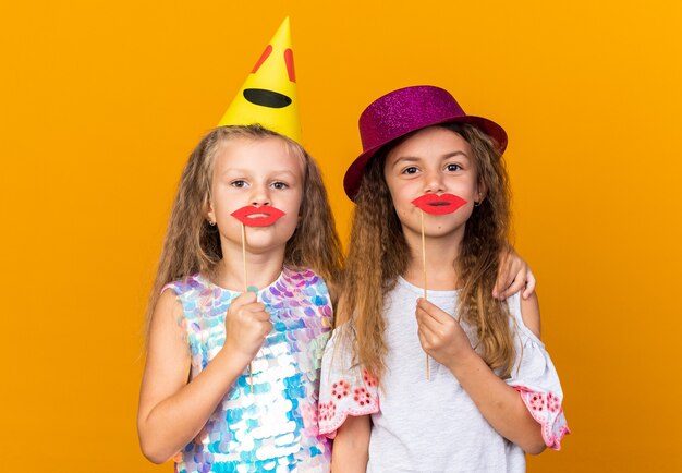 erfreute kleine hübsche Mädchen mit Partyhüten, die gefälschte Lippen auf Stöcken halten, isoliert auf oranger Wand mit Kopierraum