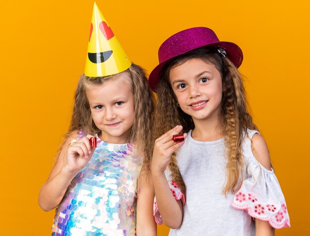 erfreute kleine hübsche Mädchen mit Partyhüten, die Partypfeifen isoliert auf oranger Wand mit Kopienraum halten