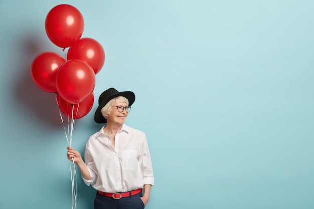 Erfreute lächelnde Dame hat faltige Haut, hat Party bei der Arbeit mit Kollegen, feiert Ruhestand, hält rote Luftballons, trägt modische Kleidung, isoliert über blaue Wand mit Leerzeichen