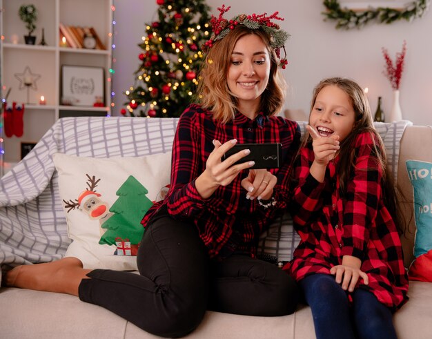 Erfreute Mutter und Tochter beobachten etwas am Telefon sitzen auf der Couch und genießen die Weihnachtszeit zu Hause