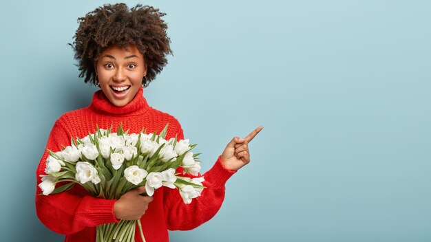 Erfreute optimistische Dame hat lockigen Haarschnitt, zeigt mit dem Zeigefinger weg, trägt roten Winterpullover, hält weiße Tulpen zeigt Leerzeichen für Ihre Werbeinhalte. Schau da! Blumen, Frauen