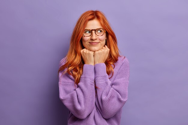 Erfreute rothaarige Frau hält Hände unter dem Kinn konzentriert beiseite hat verträumten Ausdruck trägt optische Brille und lila Pullover.