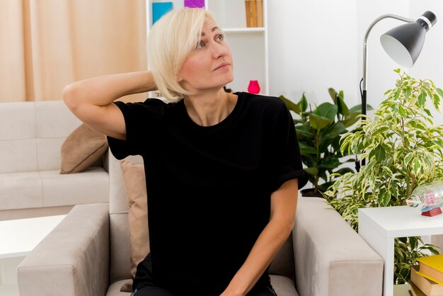Erfreute schöne blonde russische Frau sitzt auf Sessel und legt Hand auf Kopf hinter Blick auf die Seite im Wohnzimmer