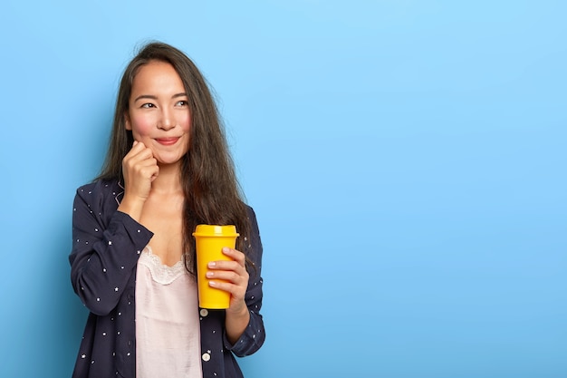 Erfreute schöne brünette asiatische Frau hat verträumten Gesichtsausdruck, überlegt, was an diesem Tag zu tun ist, wacht früh auf, trinkt erfrischten Kaffee