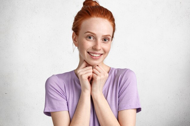 Erfreute schöne sommersprossige junge Frau mit positivem Ausdruck, lächelt freudig und freut sich über den Vorschlag eines Freundes, gekleidet in ein lässiges T-Shirt