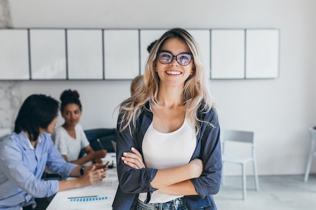 Erfreute Sekretärin in trendiger Brille posiert im Büro nach einem Treffen mit Kollegen. Innenporträt der stilvollen Geschäftsfrau mit asiatischen und afrikanischen Arbeitern.