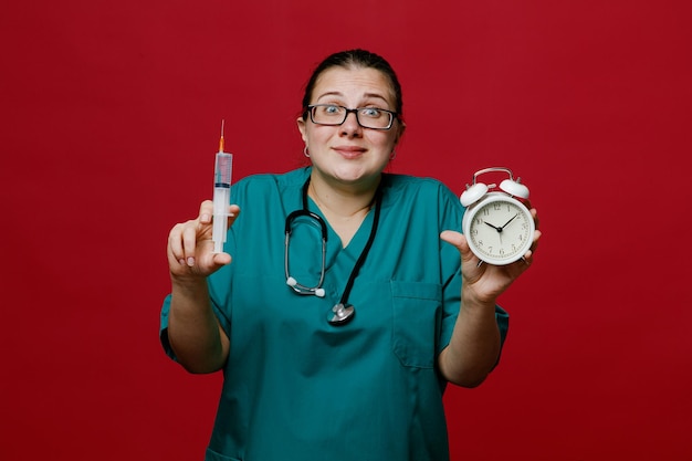 Erfreute und beeindruckte junge Ärztin, die eine einheitliche Brille und ein Stethoskop um den Hals trägt und in die Kamera blickt, die Spritze und Wecker isoliert auf rotem Hintergrund zeigt
