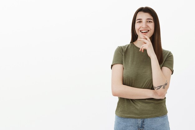 Erfreute und glückliche ehrgeizige junge Studentin mit Tätowierung und braunem Haar, das das Kinn berührt, lacht und lächelt erfreut als Gefallenidee, Denkplan ist groß stehend zufrieden über graue Wand