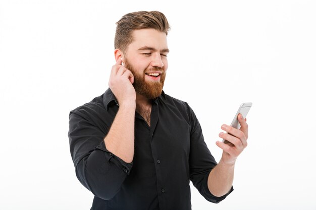 Erfreuter bärtiger Mann in hörender Musik des Hemdes durch Smartphone