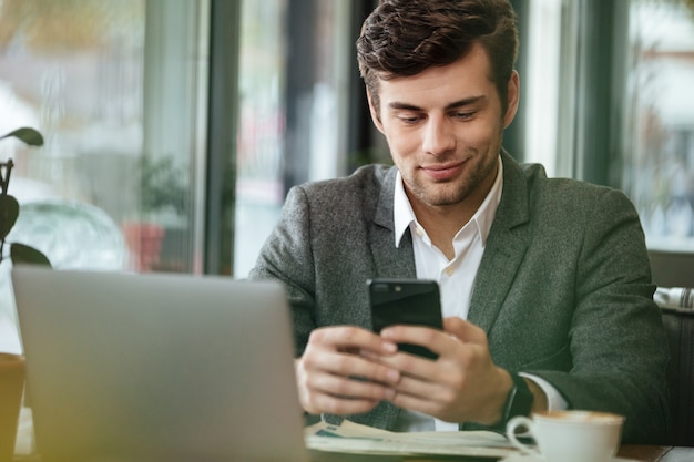 Erfreuter Geschäftsmann, der durch die Tabelle im Café mit Laptop-Computer bei der Anwendung des Smartphone sitzt