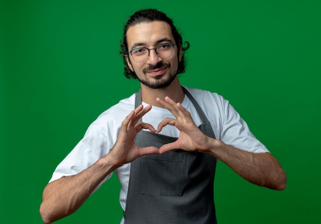 Erfreuter junger kaukasischer männlicher Friseur, der Uniform und Brille trägt, die Herzzeichen lokalisiert auf grünem Hintergrund tun