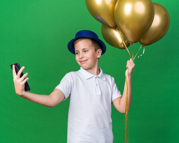 erfreuter junger slawischer Junge mit blauem Partyhut, der Heliumballons hält und selfie am Telefon einzeln auf grüner Wand mit Kopienraum nimmt