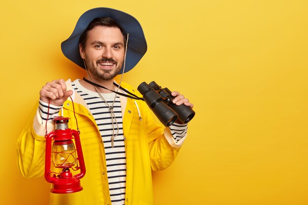 Erfreuter Mann mit Stoppeln, Kopfbedeckung und gelbem Regenmantel, Petroleumlampe und Fernglas, fröhlich in die Kamera schauend, drinnen stehend