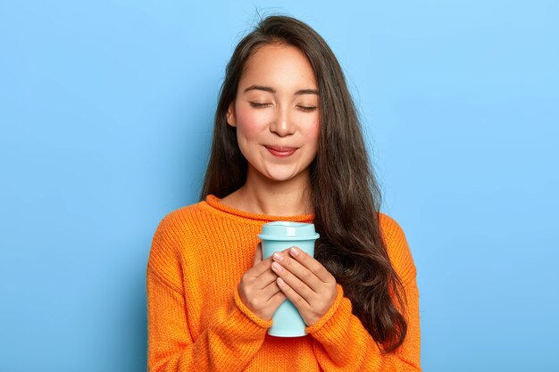 Erfreutes erholsames Mädchen mit asiatischem Aussehen, hält die Augen geschlossen, lächelt sanft, trinkt gerne aromatischen Espresso aus der Tasse zum Mitnehmen und trägt einen orangefarbenen Pullover