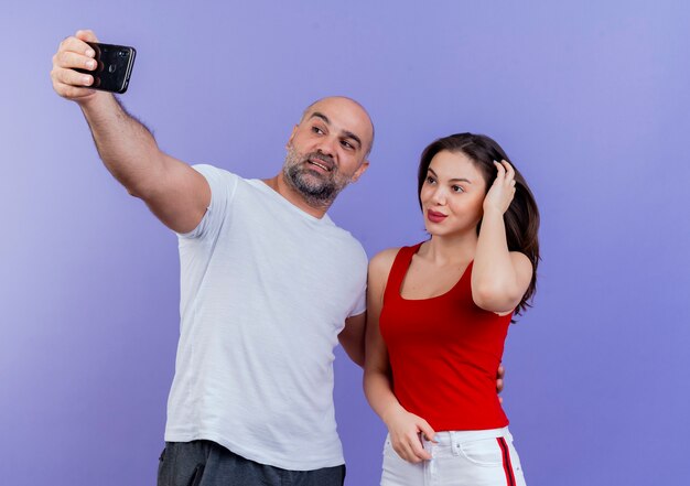 Erfreutes erwachsenes Paar, das Selfie-Mann nimmt, der Hand auf die Taille der Frau legt und sie den Kopf berührt